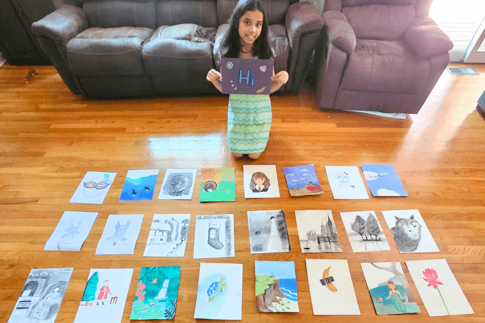 Young artist Uma smiling and holding her artworks from Nimmy's Art Online Art Classes and surrounded by over 20 colorful artworks displayed neatly in rows.  