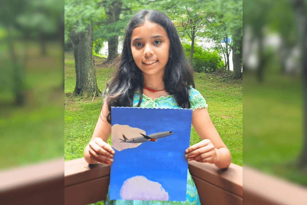 Young artist Uma holding a gouache painting of a passenger airplane flying through a bright blue sky with clouds, while standing outdoors.  