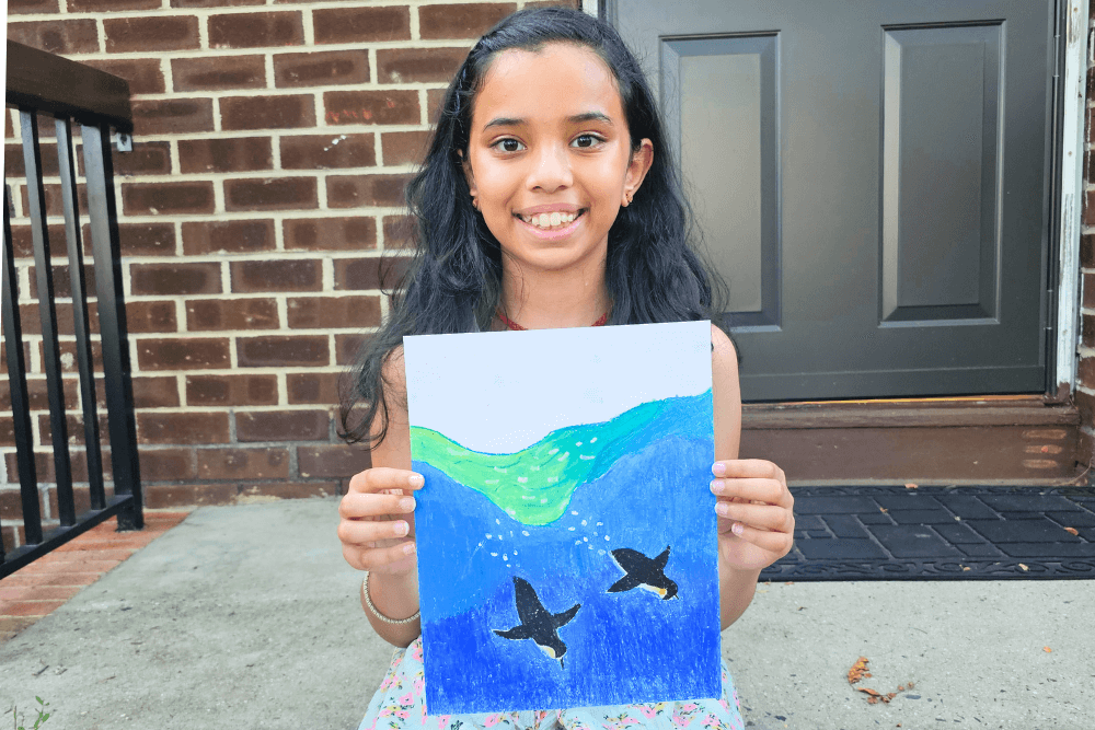 Young artist Uma smiling and holding an oil pastel drawing of two penguins swimming underwater, with vibrant blue and green tones.