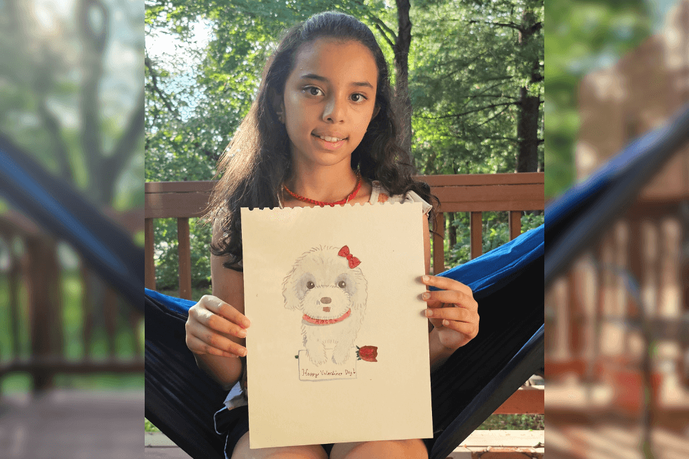 Young artist Uma holding a Valentine-themed watercolor painting of a fluffy white dog with a red bow and rose, with the words “Happy Valentine’s Day.”