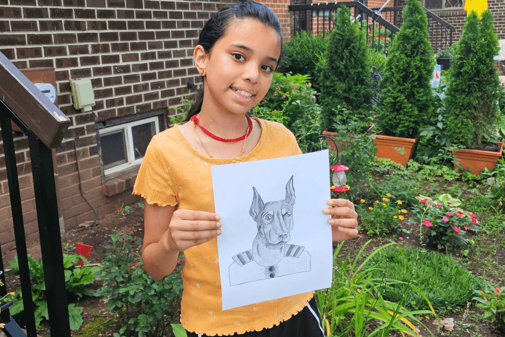 Young artist Uma smiling and holding a charcoal drawing of a regal Doberman wearing a military-style uniform.