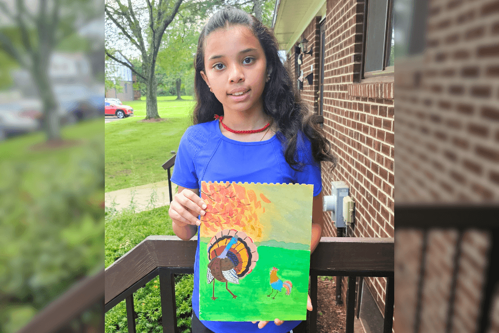 Young artist Uma standing outdoors, holding a Thanksgiving-themed acrylic painting of a turkey and rooster beneath an autumn tree with orange leaves.
