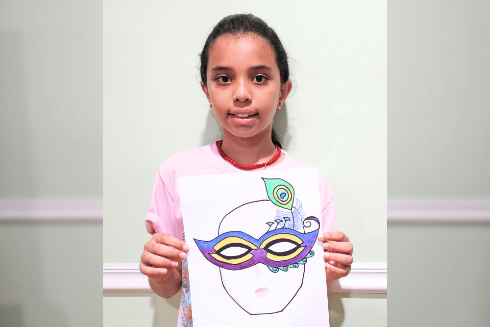 Young artist Uma holding a colored pencil drawing of a Venetian-style mask with decorative feathers and bold colors, drawn on a stylized face outline.