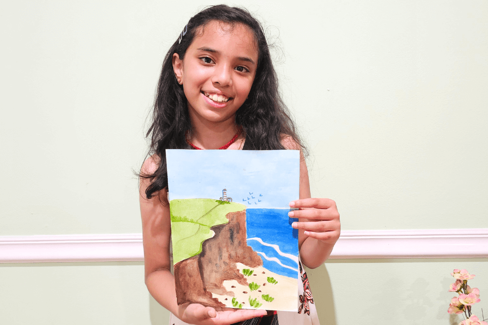Young artist Uma smiling and holding a watercolor painting of a dramatic cliffside meeting the ocean, complete with a lighthouse, beach, and flying birds.