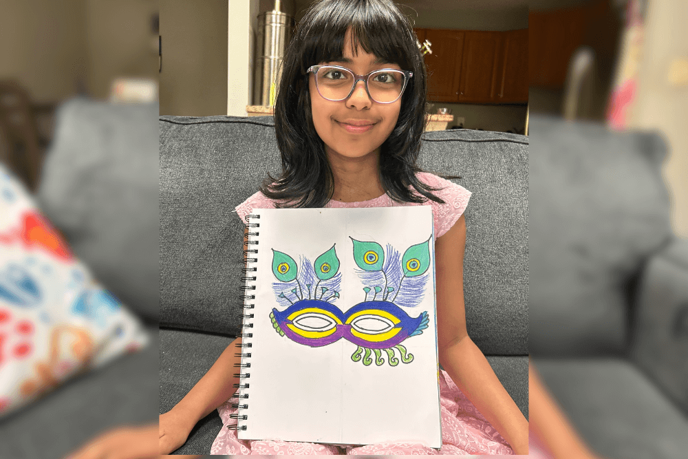 Aadhya holding her Prismacolor drawing of a Venetian eye mask with peacock feathers, exploring symmetry and asymmetry.