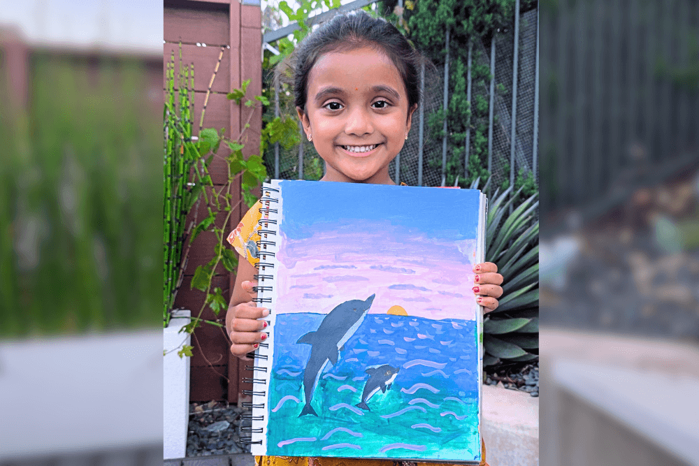 Aanya holding her acrylic painting Dolphins’ Surfing Time showing playful dolphins jumping in the ocean at sunset.