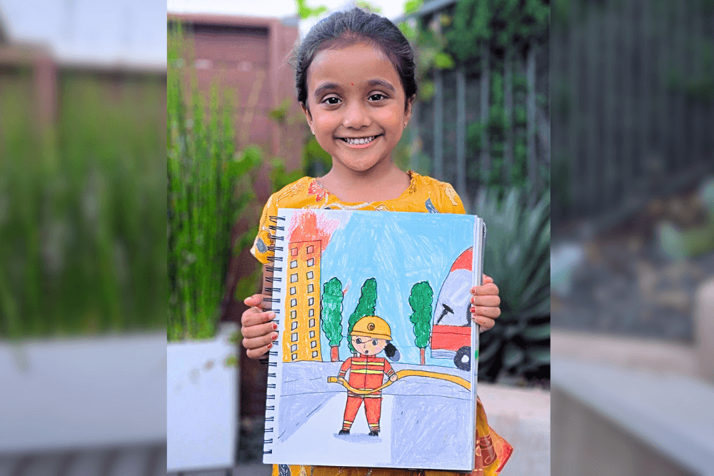 Aanya holding her oil pastel drawing of a firefighter and fire truck in a city scene.