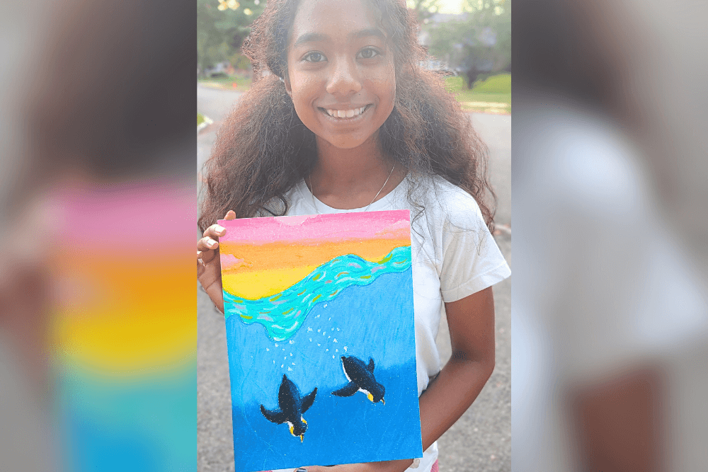 Harene holding her oil pastel artwork of penguins swimming, created using analogous colors.