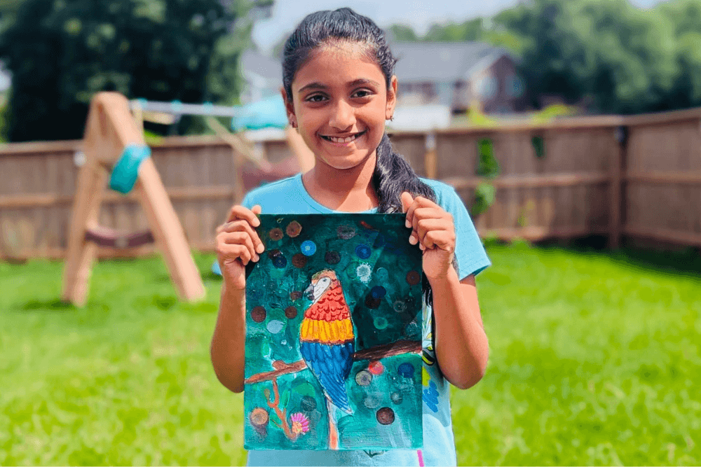 Joann holding her colorful gouache painting Colors of the Macaw with bright feathers and playful details. From summer art camp.