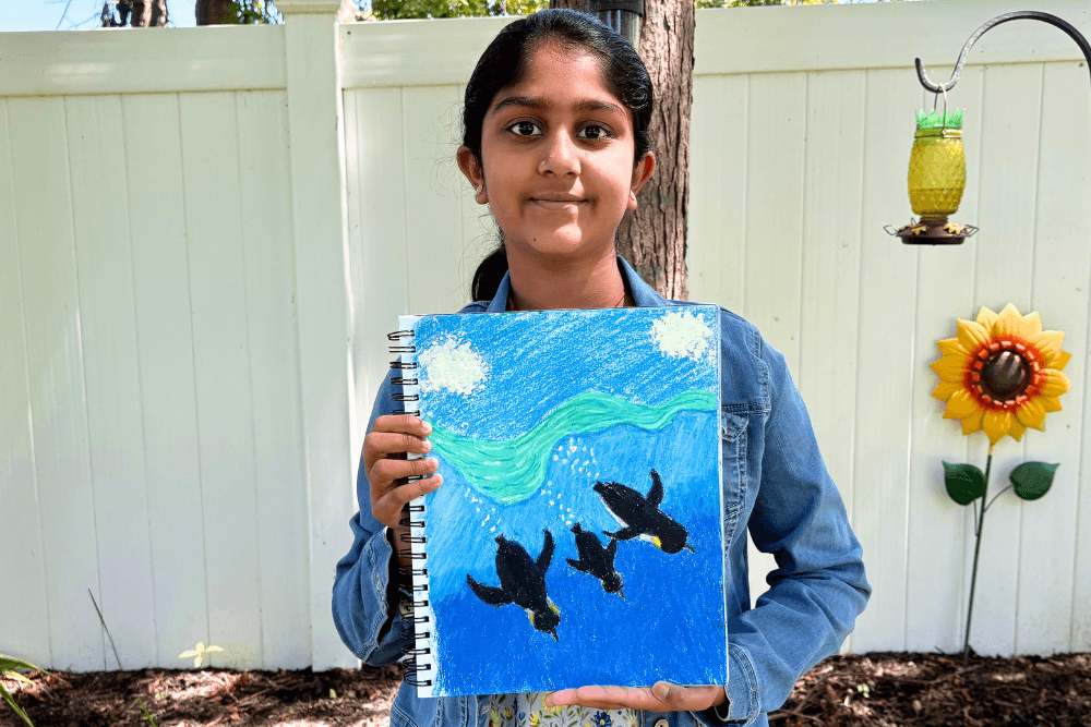 Navya holding her oil pastel painting of three penguins swimming under a bright blue sky.