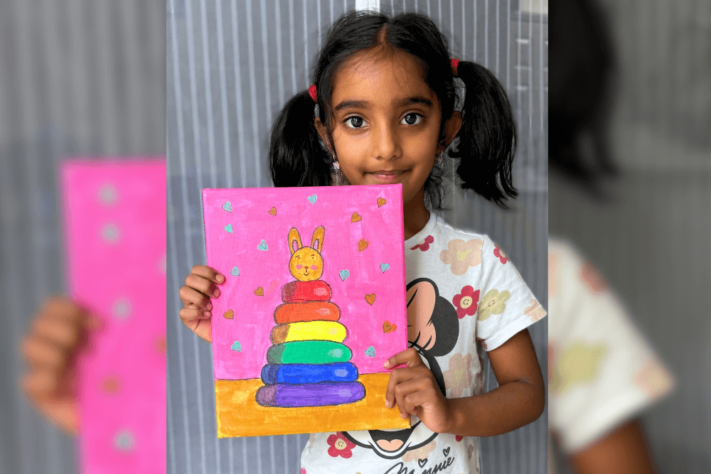 Pranavi holding her acrylic painting of a rainbow stacking toy still life with a colorful pink and gold background.