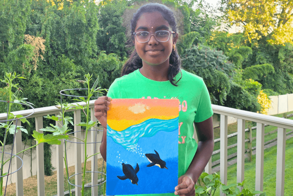 Sananda holding her oil pastel painting Penguins, created with analogous colors, showing penguins swimming under a bright sunset.