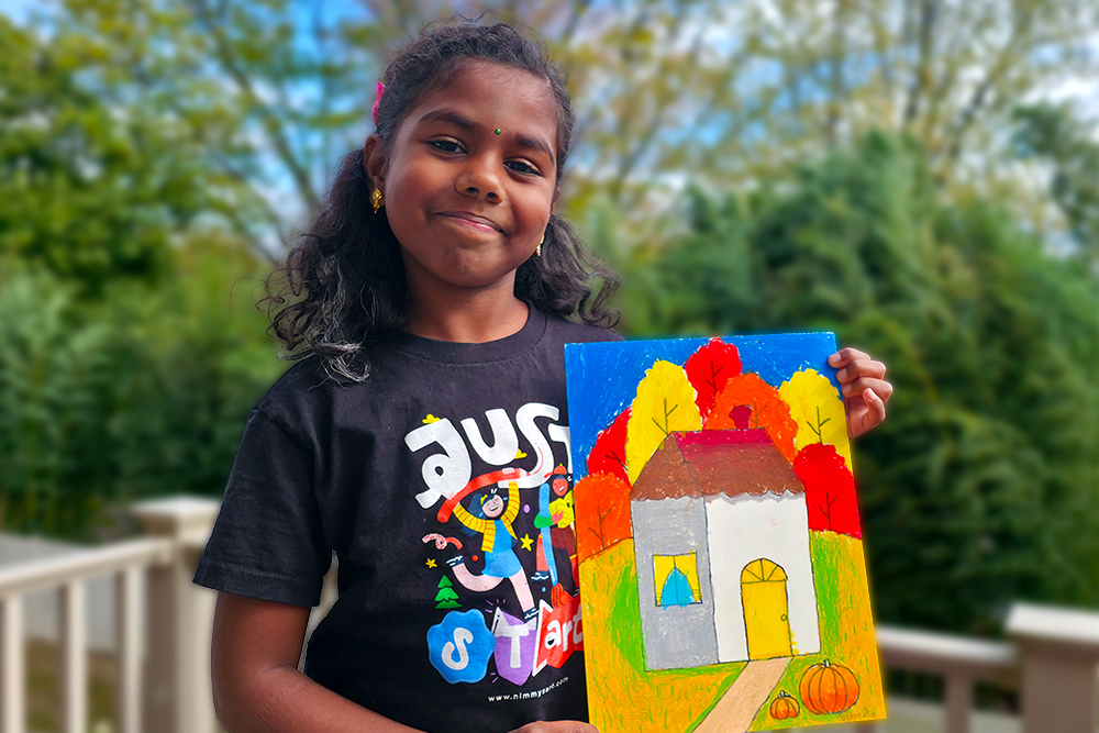 Sanmita proudly holds her fall-themed painting, featuring a cozy white house surrounded by colorful autumn trees, golden leaves, and a pumpkin on the path.