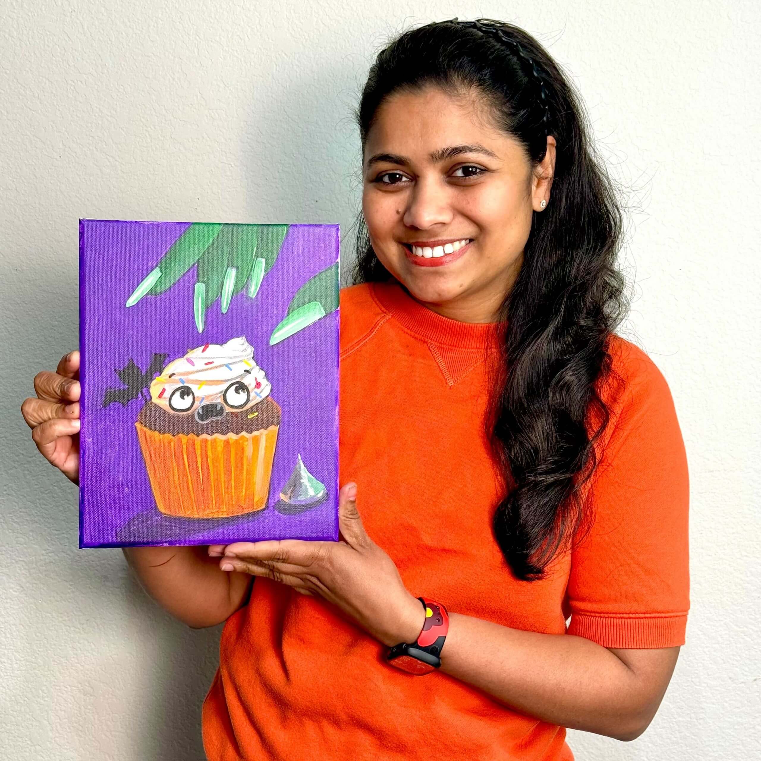 Art instructor holding a “Spooky Cupcake” Halloween painting for Nimmy’s Art Halloween Art Contest 2025 for young artists.
