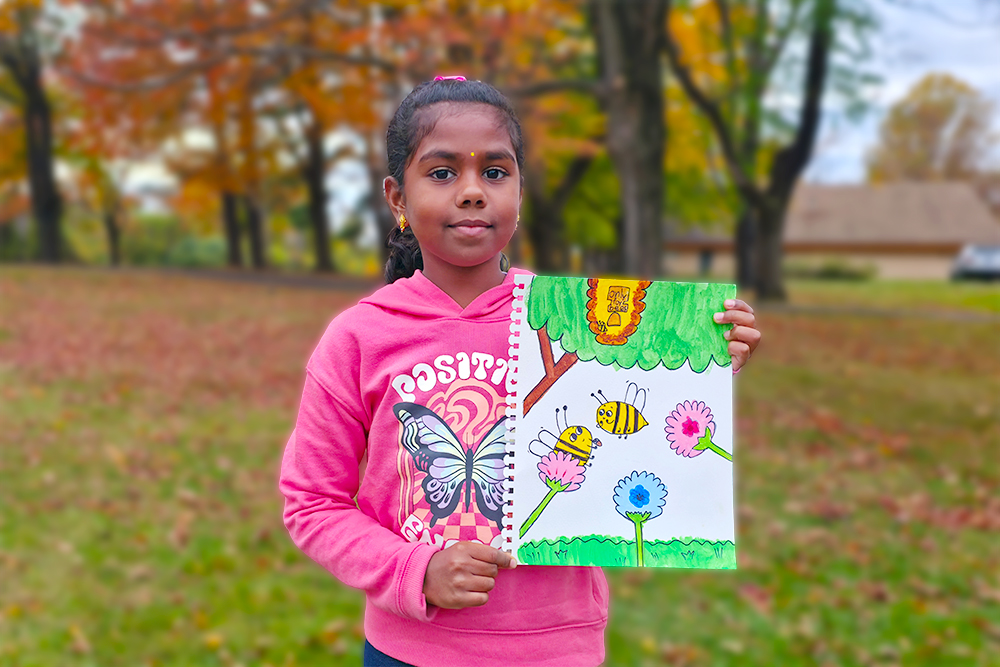 Sanmita, a 7-year-old young artist, holds her cheerful acrylic painting titled “World Bee Day.” Her artwork features two happy bees buzzing around colorful flowers under a bright, sunny sky, spreading an inspiring message about the beauty of nature and the importance of protecting bees.