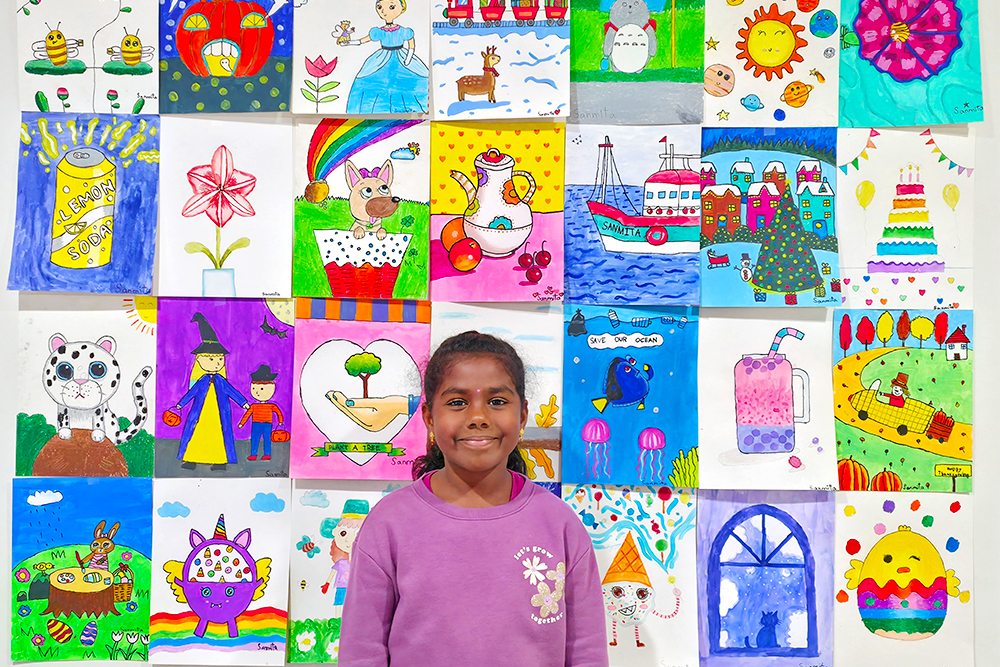 Sanmita, a 7-year-old young artist, stands proudly in front of her colorful art wall displaying all her creative paintings. Her artwork features bright and cheerful subjects like animals, landscapes, and imaginative characters, showing her love for art and storytelling.