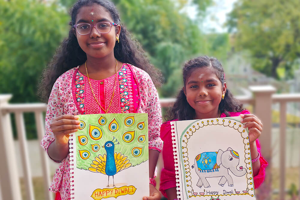 Sanmita proudly holds her Diwali-themed artwork titled “Jumbo Circus,” a charming pencil and sharpie drawing of a festive elephant, while her sister Sananda also holds her colorful “Peacock in Abstract Colors” painting. Both sisters smile brightly, celebrating the spirit of Diwali through their creative art.