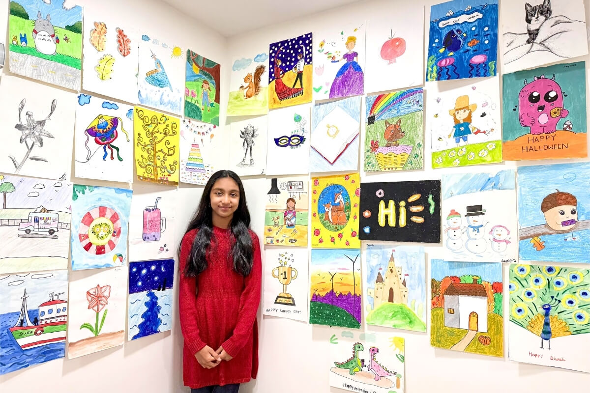 Aayushi, an 8-year-old young artist, stands proudly in front of her vibrant art wall showcasing a collection of her creative artworks. Her pieces feature a wonderful mix of colorful scenes, imaginative characters, landscapes, and playful designs, reflecting her growing artistic skills and her love for expressing stories and ideas through art.