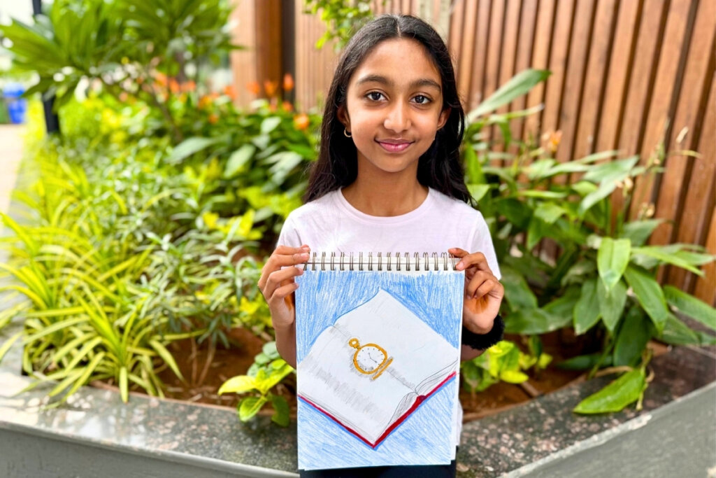 Aayushi proudly holds her Prismacolor colored pencil artwork, a still life study of a golden pocket watch resting on an open book, carefully shaded with soft blues and detailed textures.