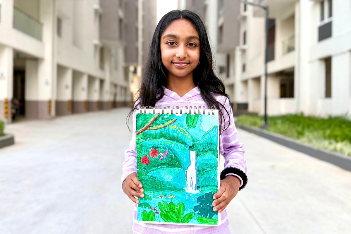 Aayushi proudly holds her gouache painting “Rainforest,” featuring lush green trees, layered foliage, a flowing waterfall, and colorful details inspired by nature.