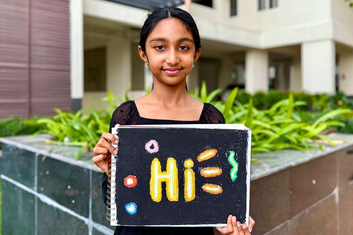 Aayushi proudly holds her oil pastel artwork “Neon Sign Board,” featuring the word “Hi” glowing in bright neon colors against a dark background, highlighting strong contrast and value.
