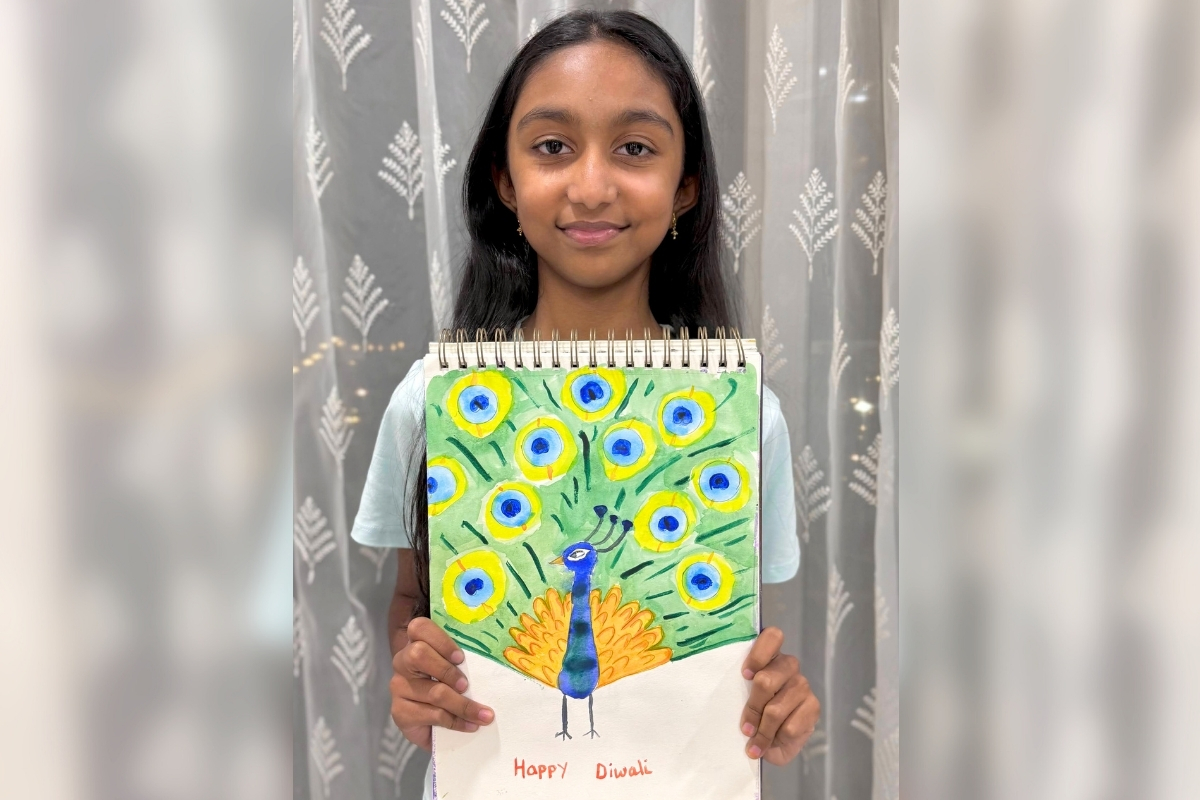 Young artist Aayushi smiling while holding her watercolor painting of a peacock in abstract colors, created as a Diwali-themed artwork, featuring vibrant blues, greens, and festive details.