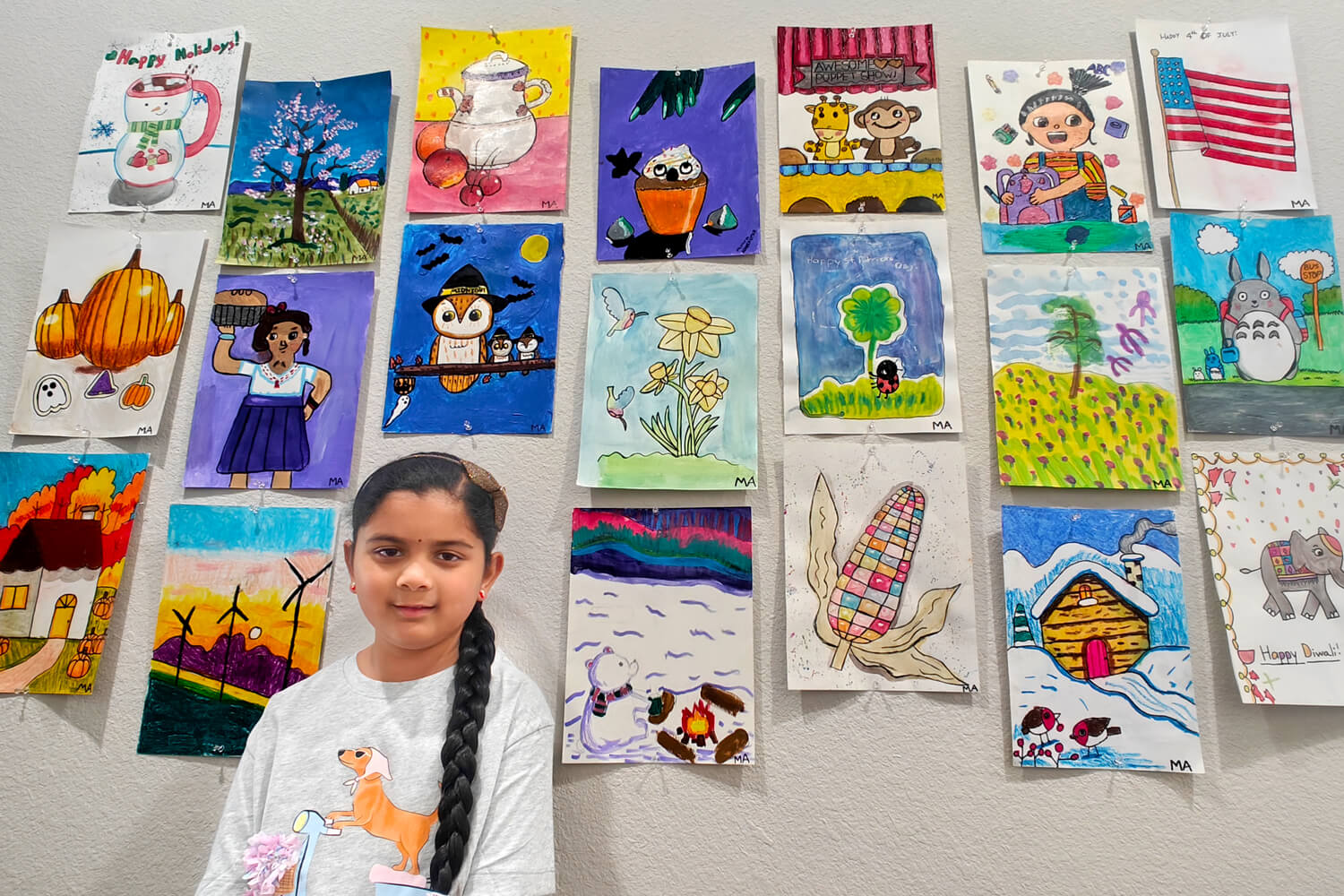 Mithra, a 7-year-old girl, stands smiling in front of a wall filled with her colorful paintings.