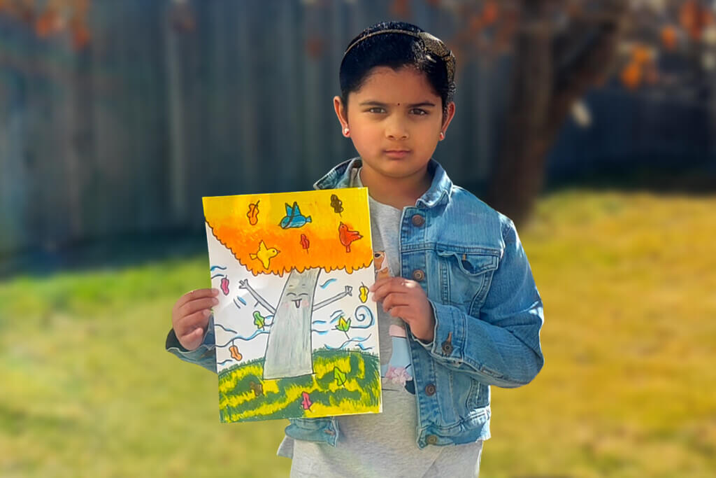 Mithra, a 7-year-old girl, stands outdoors holding her oil pastel artwork of a fall tree with colorful leaves.