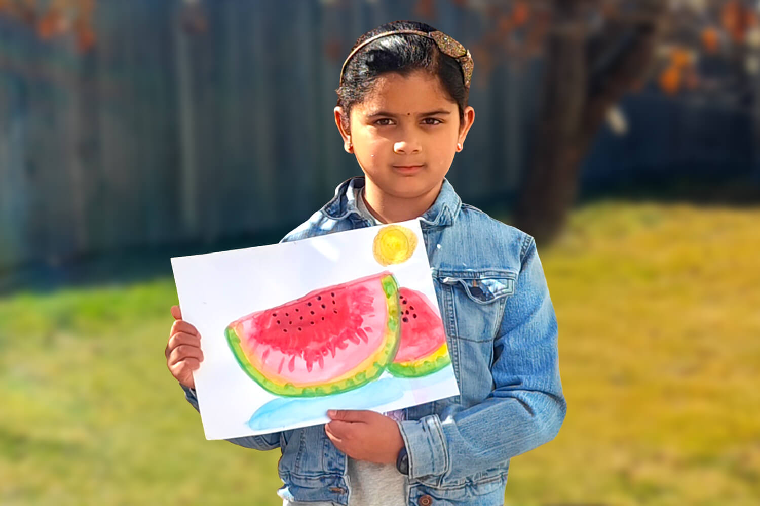 Mithra, a 7-year-old girl, stands outdoors holding her watercolor painting of a bright red watermelon slice, looking proudly at the camera.