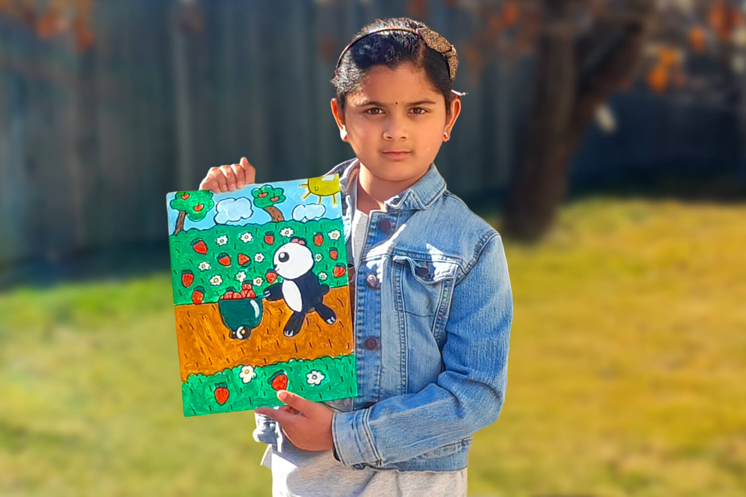 Mithra, a 7-year-old girl, stands outdoors holding her acrylic painting of a panda picking strawberries in a bright and colorful field.