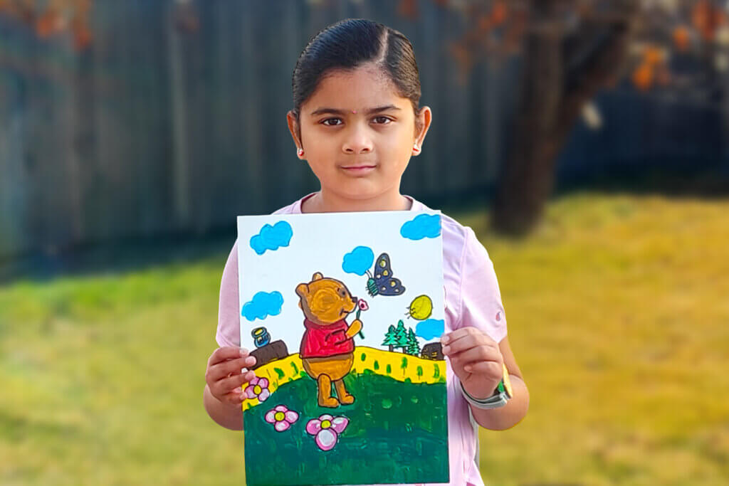 Young girl named Mithra holding her colorful acrylic painting of Winnie the Pooh standing in a bright outdoor scene with flowers, trees, clouds, and a butterfly.