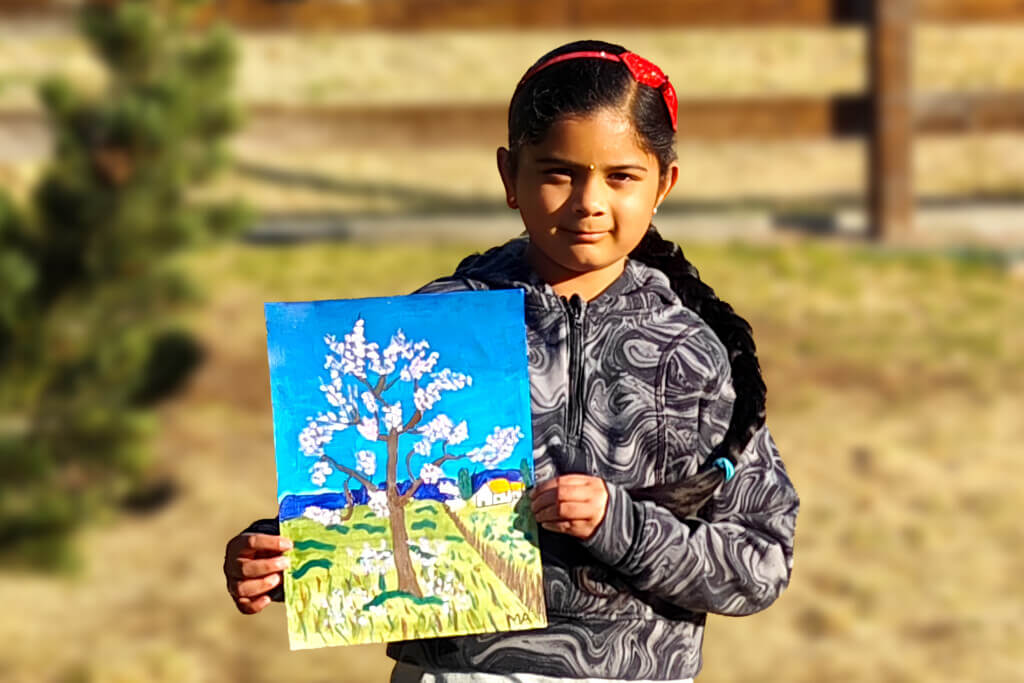 Young artist Mithra holding her acrylic painting of a blooming peach tree inspired by a Van Gogh master study, featuring a bright blue sky, green fields, and a small house in the background.