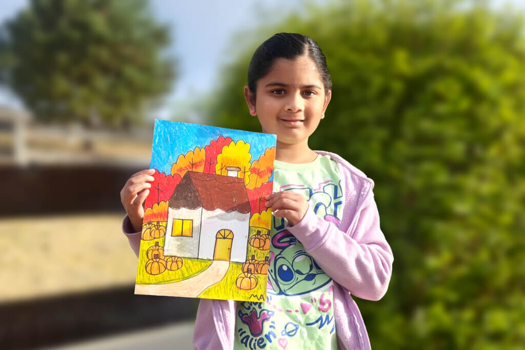 Mithra, a 7-year-old girl, stands outdoors holding her oil pastel painting of a small house with colorful autumn trees and pumpkins.