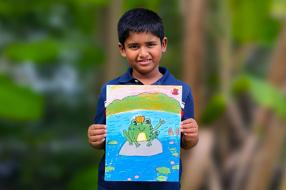 Sarthak, a young artist, proudly holds his oil pastel painting titled “Cute Froggy.” The artwork features a cheerful green frog wearing a crown and sitting on a lily pad surrounded by bright blue water, blooming water lilies, and a peaceful pond landscape. With vibrant colors and playful details, the painting captures the charm of nature and showcases Sarthak’s creativity and growing artistic skills.