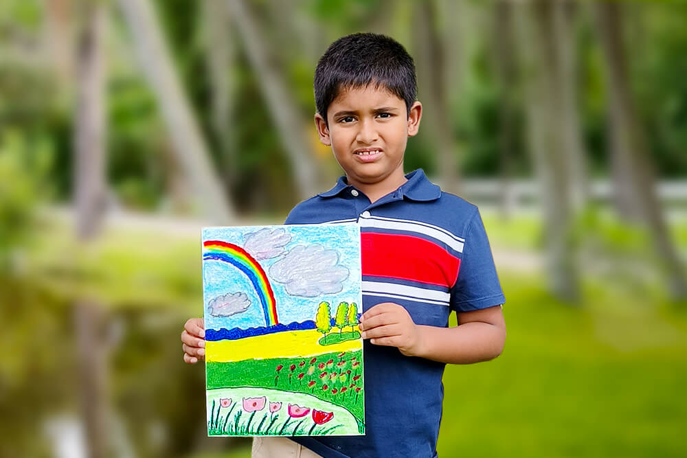 Sarthak, a young artist, proudly holds his oil pastel painting titled “Colors of Nature.” The artwork features a bright rainbow stretching across the sky above a colorful landscape filled with green fields, blooming flowers, fluffy clouds, and trees, celebrating the beauty of the natural world.