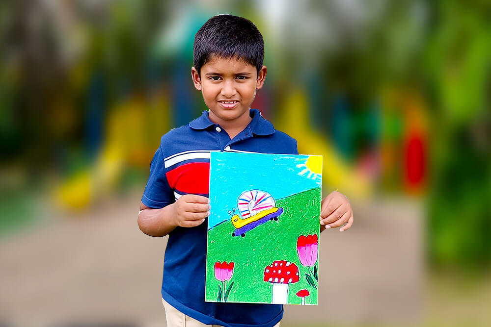 Sarthak, a young artist, proudly holds his oil pastel painting titled “Skating Snail.” The artwork features a cheerful snail riding a skateboard across a grassy hill, surrounded by colorful flowers, a bright sun, and whimsical mushrooms in a playful outdoor scene.