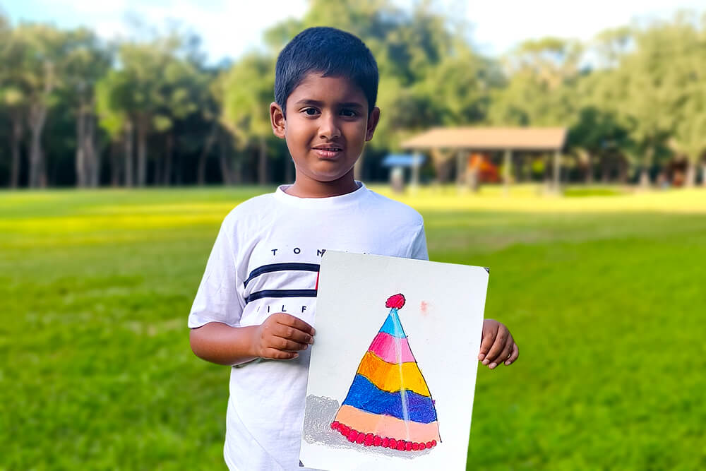 Sarthak, a young artist, proudly holds his oil pastel painting titled “Colorful Birthday Hat.” The artwork features a festive party hat decorated with bright rainbow-colored stripes and playful details, creating a cheerful and celebratory design.