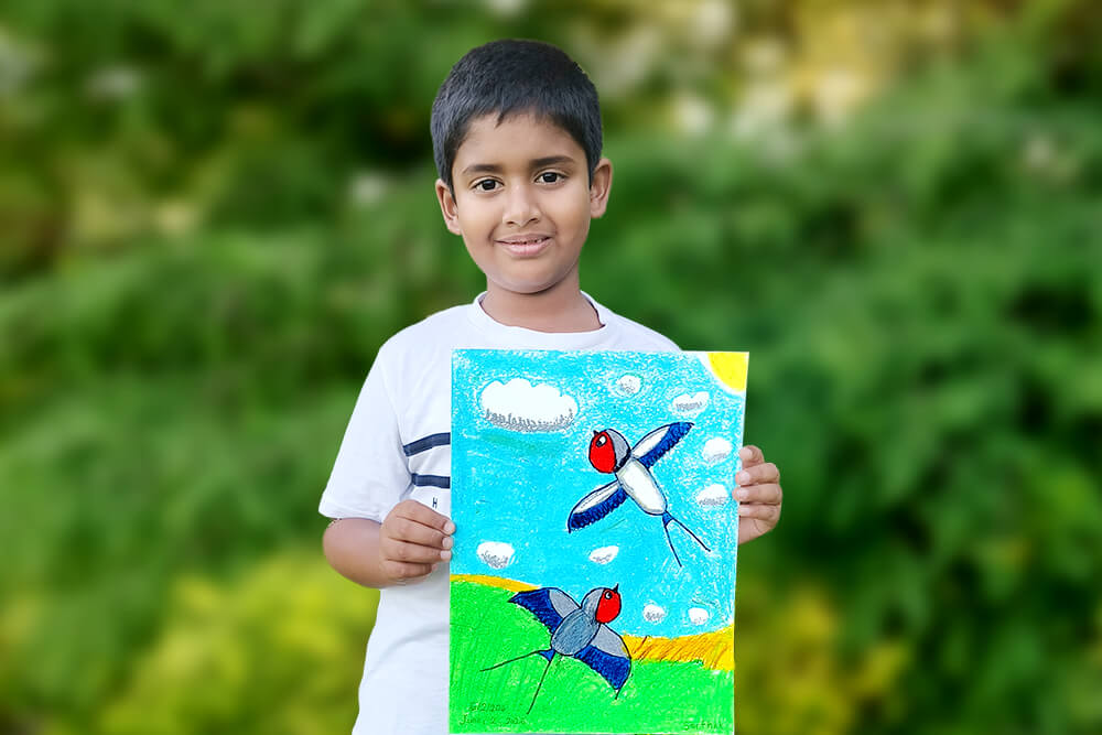 Sarthak, a young artist, proudly holds his oil pastel painting titled “Dancing Swallows.” The artwork features two colorful swallows soaring through a bright blue sky above green fields, capturing the grace and movement of birds in flight.