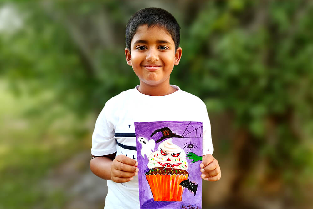 Sarthak, a young artist, proudly holds his acrylic painting titled “Halloween Spooky Cupcake.” The artwork features a spooky cupcake decorated with a creepy face, a witch’s hat, a ghost, a spider, and a bat, creating a fun Halloween-themed scene filled with colorful details.