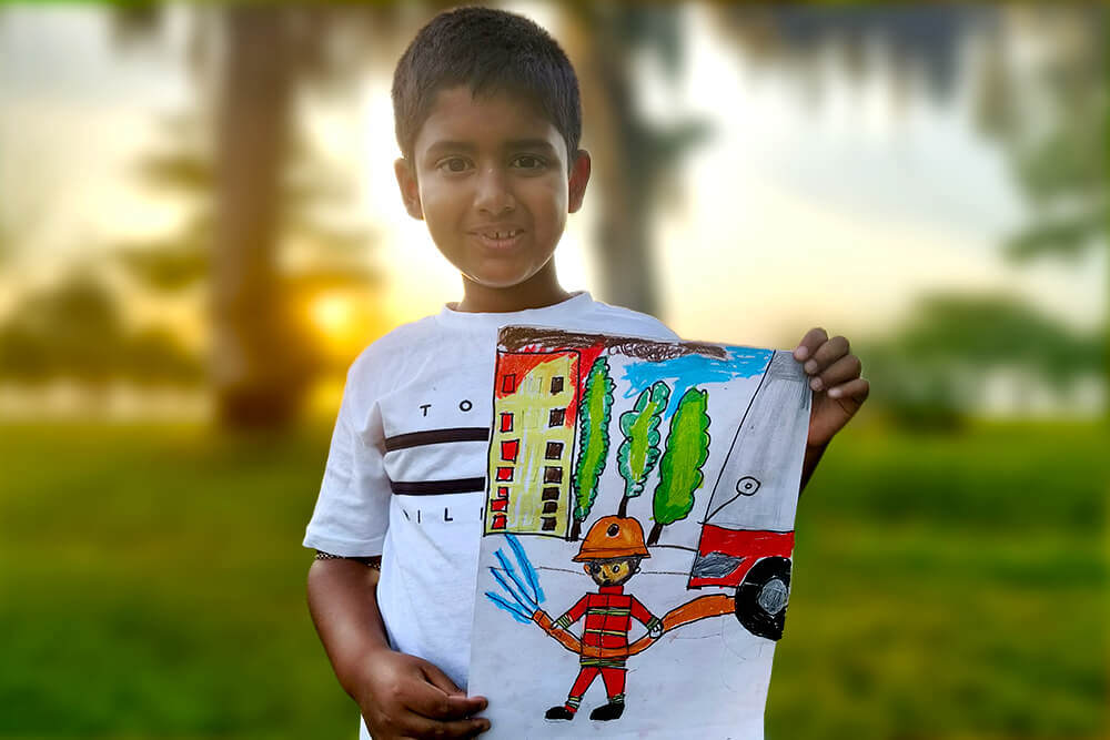 Sarthak, a young artist, proudly holds his oil pastel painting titled “Fire Truck on the Way.” The artwork features a firefighter spraying water from a hose beside a fire truck, with buildings and trees in the background, capturing the bravery and action of firefighters at work.