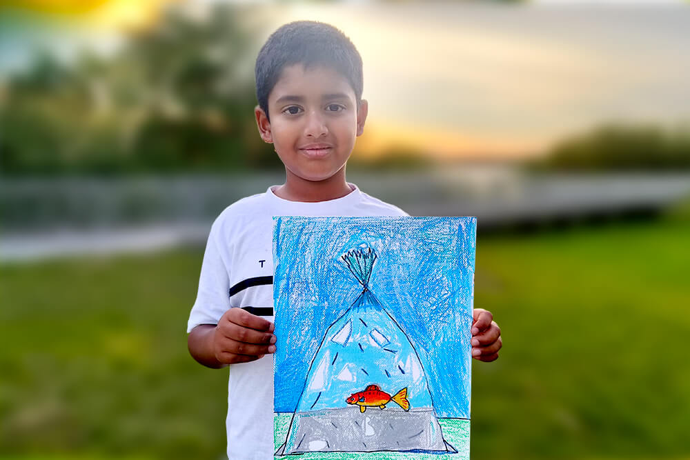 Sarthak, a young artist, proudly holds his oil pastel painting titled “Goldfish in a Bag.” The artwork features a bright orange goldfish swimming inside a clear water-filled bag, with shades of blue and reflective details that create a playful and realistic underwater effect.