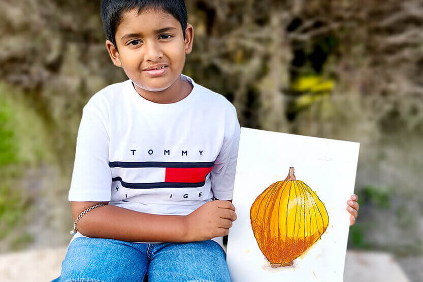 Sarthak, a young artist, proudly holds his oil pastel painting titled “3D Pumpkin.” The artwork features a bright orange pumpkin with realistic shading and highlights, creating a three-dimensional effect that makes the pumpkin appear to pop off the page.
