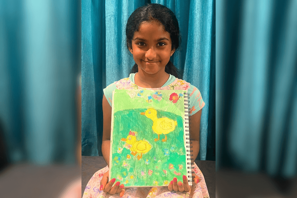 Ameya Kolachina holding her artwork of two yellow ducks surrounded by colorful flowers in a green meadow.
