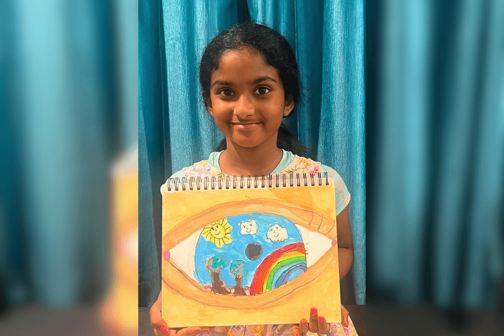 Ameya Kolachina holding her acrylic eye painting with a rainbow, trees, smiling sun, and clouds inside the blue iris.