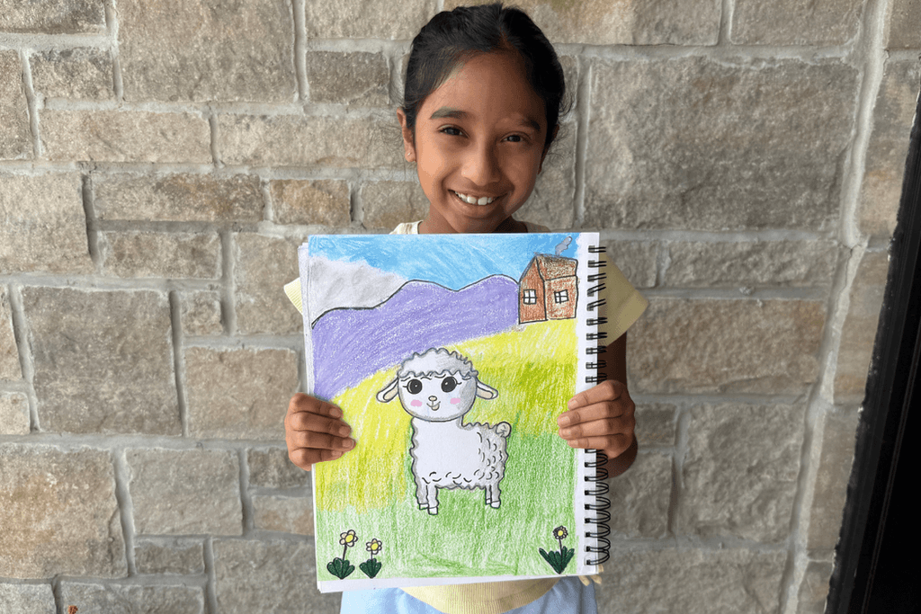 Ira Nadella holding her Baby Lamb oil pastel artwork with a white lamb, green meadow, purple mountains, and a small house.