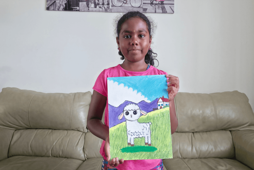 Sanmita holding her Baby Lamb oil pastel artwork, showing a white lamb in a green field with purple mountains and a house.