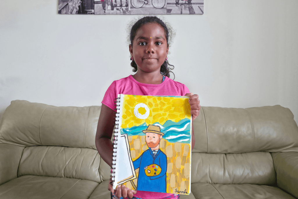 Sanmita holding her acrylic painting of Vincent van Gogh in a straw hat and blue coat, with a palette beside an easel.