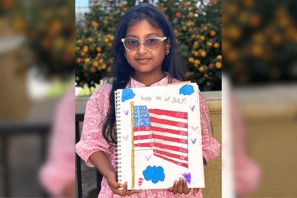 Shloka holding her Fourth of July drawing of an American flag surrounded by colorful hearts and blue clouds.