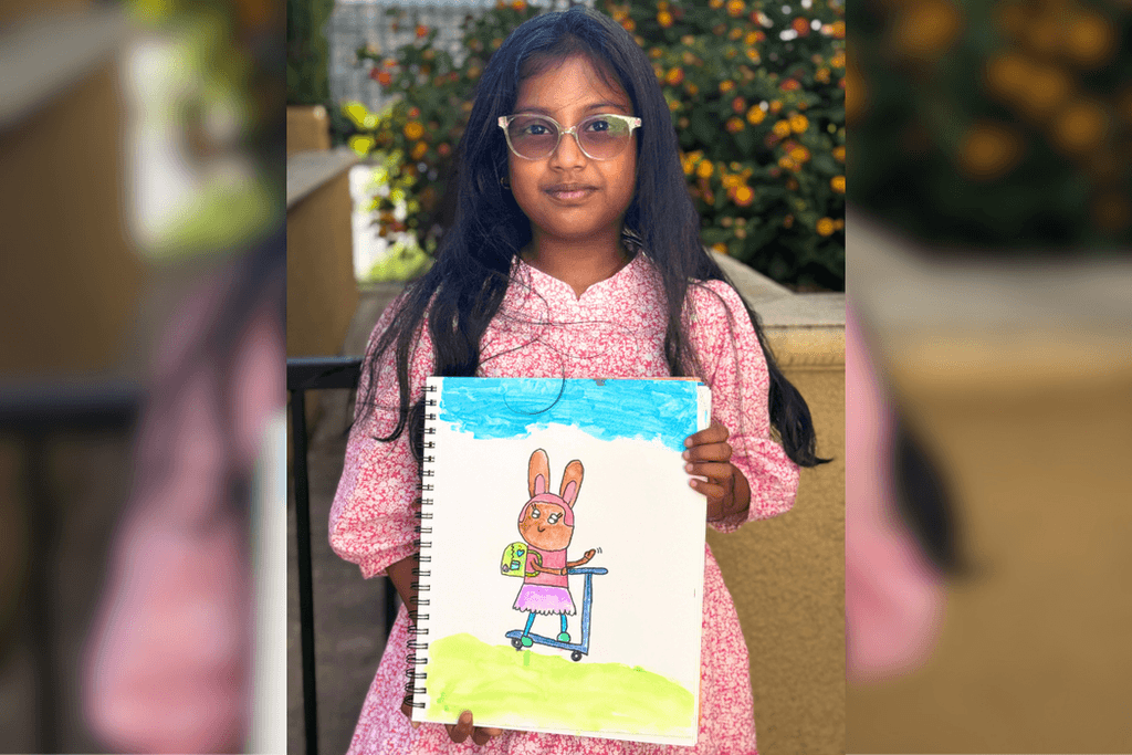 Shloka holding her drawing of a rabbit wearing a backpack and riding a blue scooter beneath a blue sky.