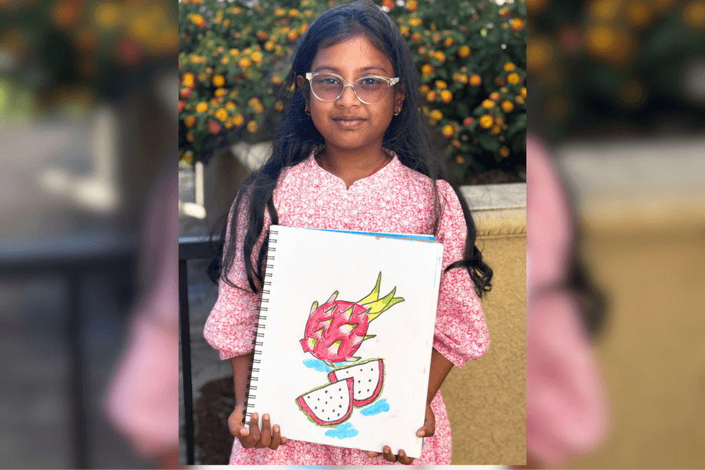 Shloka holding her dragon fruit drawing with bright pink skin, green tips, and two white slices dotted with black seeds.
